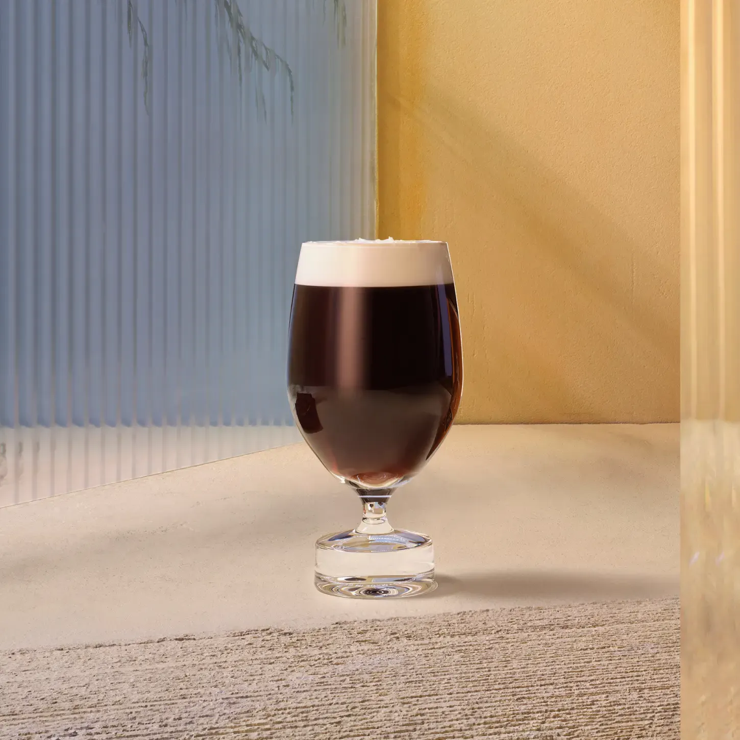 A glass of dark coffee with a creamy foam top sits on a textured surface. Its surrounded by softly lit beige and translucent panels, creating a warm and minimalist setting.