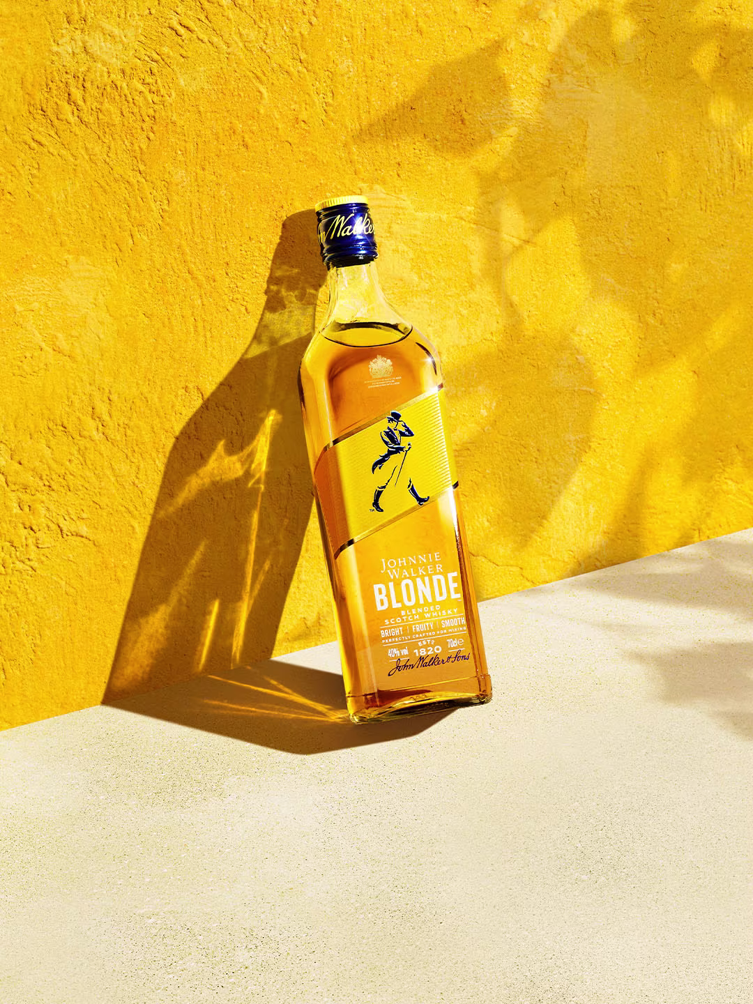 Butelka whisky Johnnie Walker Blonde stoi pionowo na tle jaskrawożółtej ściany, rzucając cień na jasną powierzchnię, a światło słoneczne tworzy ciepłą i letnią atmosferę.