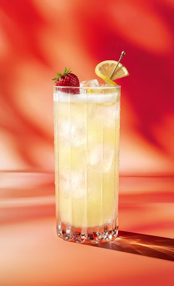 Ein hoher, eisgekühlter Cocktail, garniert mit einer Zitronenscheibe und einer Erdbeere, vor einem orange-roten Hintergrund.