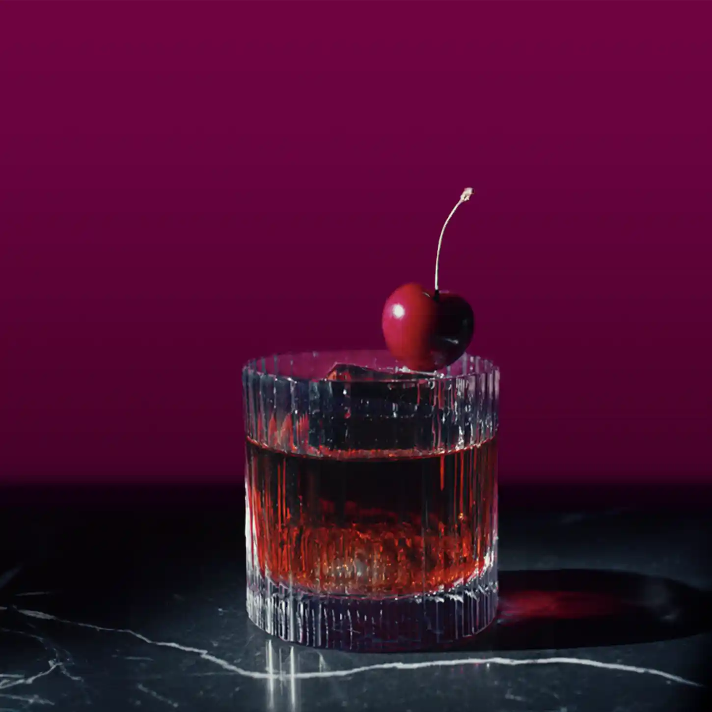 A dark red cocktail glass sits on a marble surface against a dark brown background. The drink is garnished with a cherry on the rim. The glass casts a shadow to the right, indicating a light source from the left.