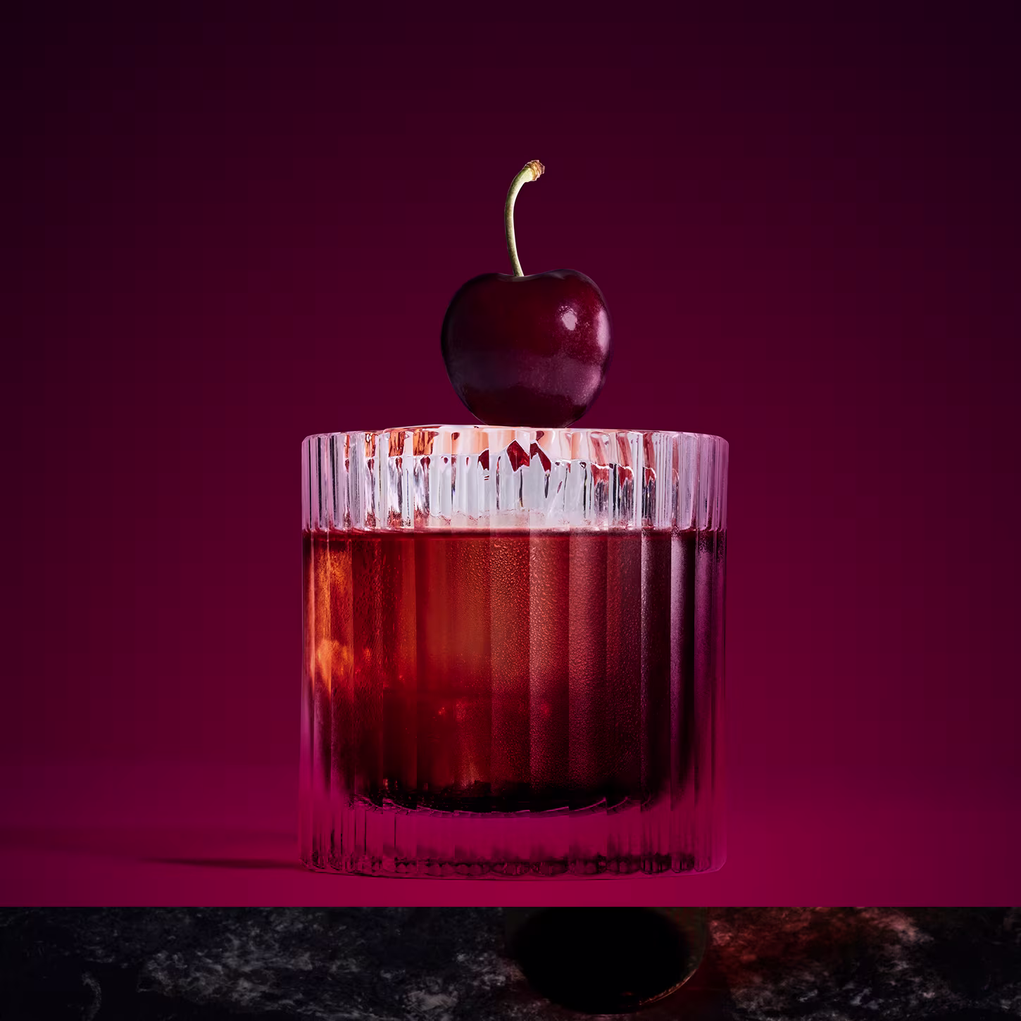 Una copa de cóctel rojo oscuro con hielo, decorada con una cereza sobre un fondo magenta intenso. La copa reposa sobre una superficie de mármol oscuro.