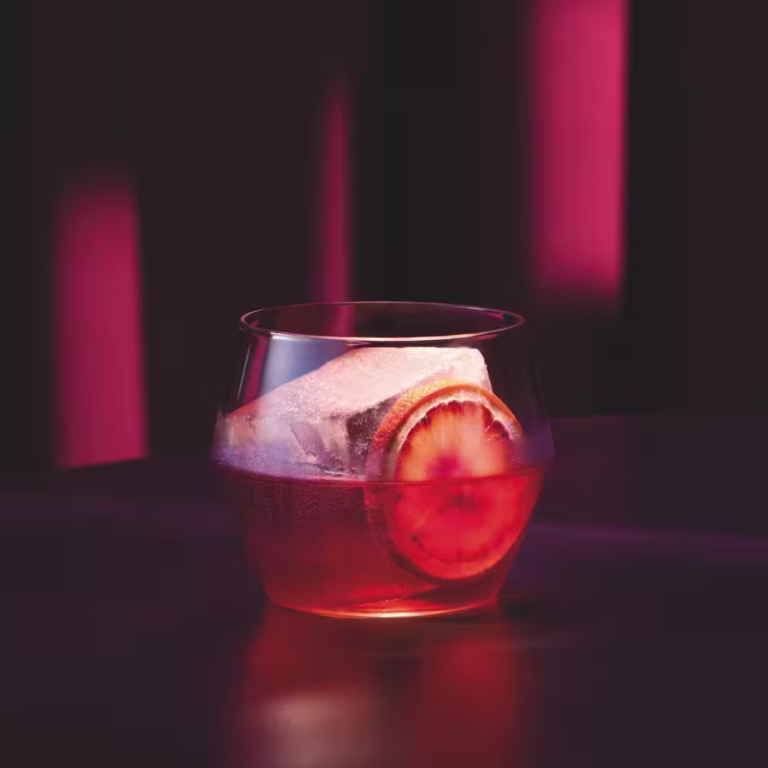 A glass of red cocktail with a large ice cube and an orange slice sits on a dark surface, with dramatic pink and purple lighting in the background.