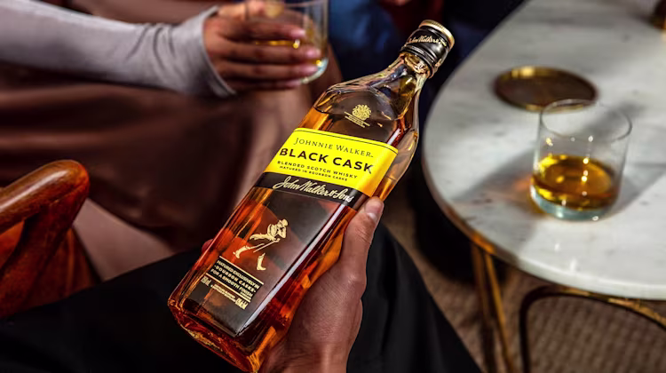 A person holds a bottle of Johnnie Walker Black Cask whisky, with two glasses of whisky on a white marble table nearby. Another person holds a drink in the background.