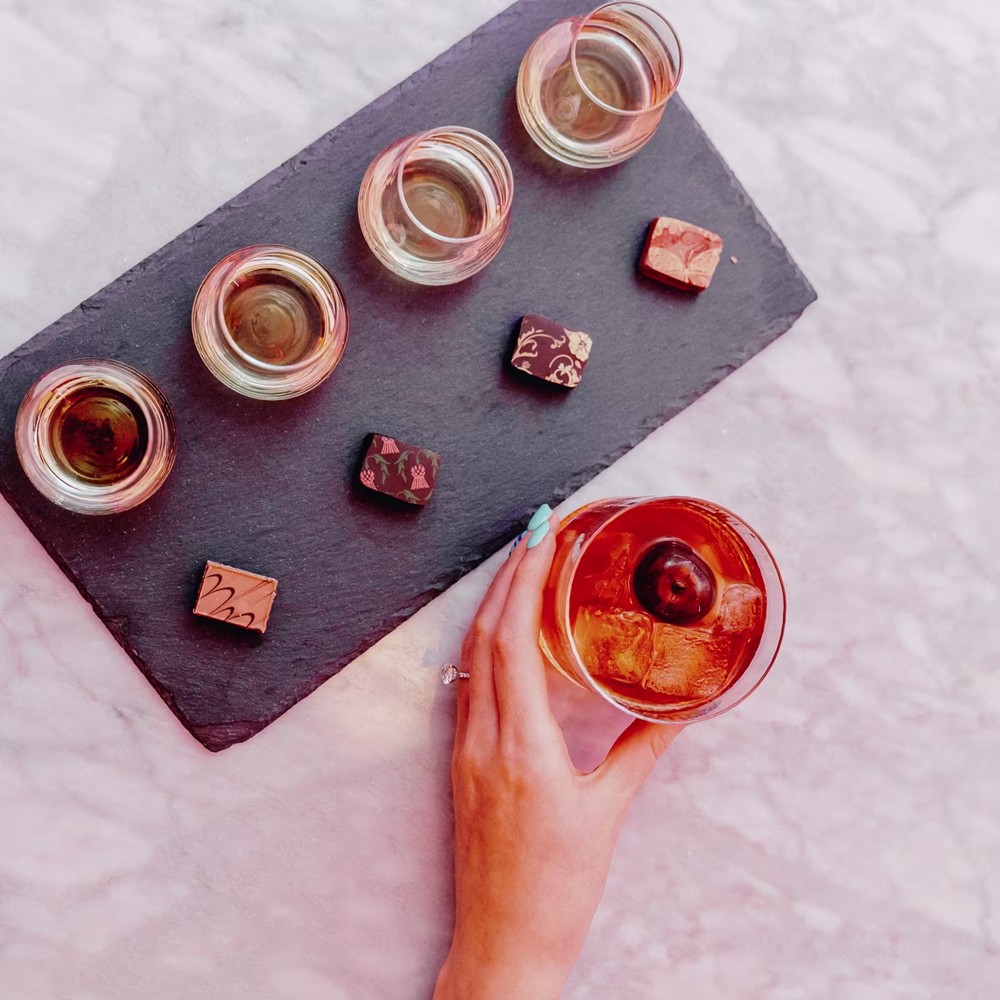 A hand holds a cocktail beside four whiskey glasses and four assorted chocolates on a slate tray.