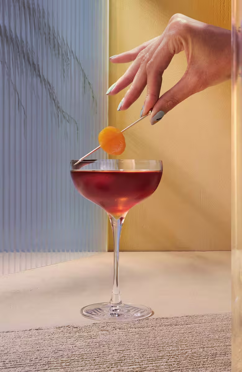 A manicured hand places an orange garnish on a cocktail in a coupe glass, set against a beige and blue textured background.