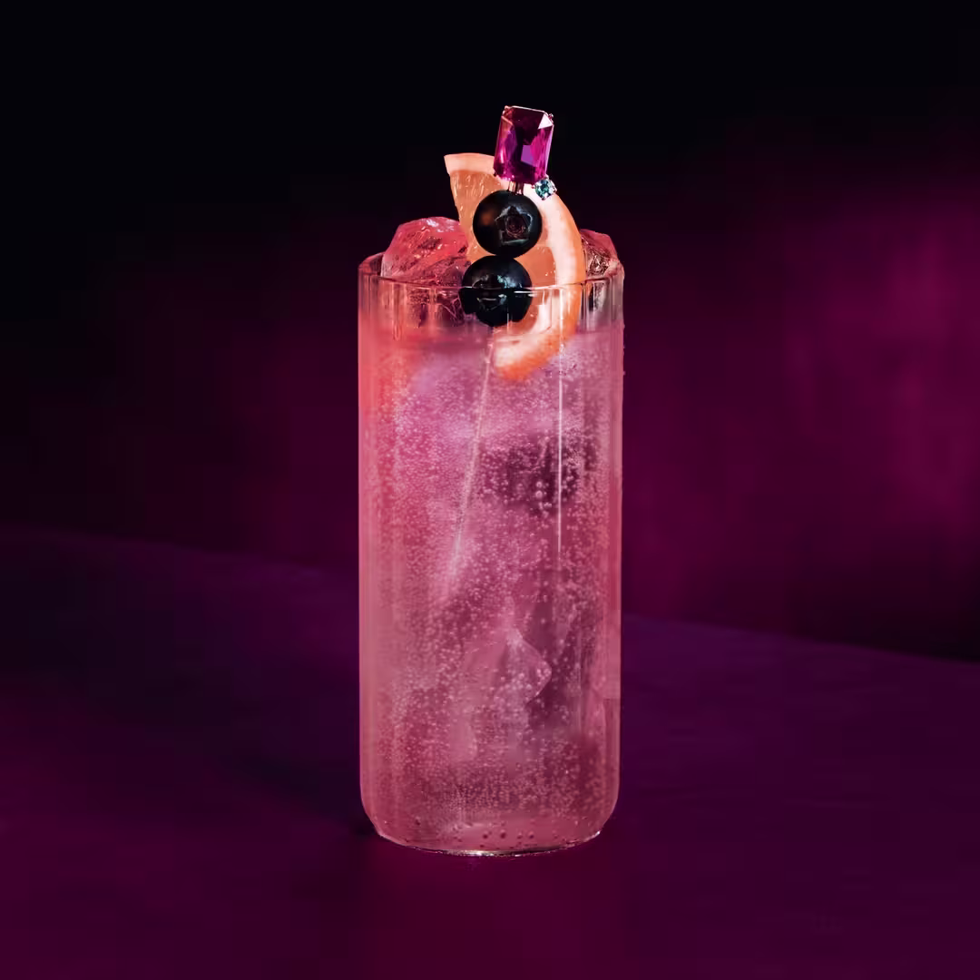 A tall glass filled with a sparkling pink cocktail, ice cubes, and garnished with two dark cherries and a grapefruit slice, sits on a magenta surface against a dark, gradient background.