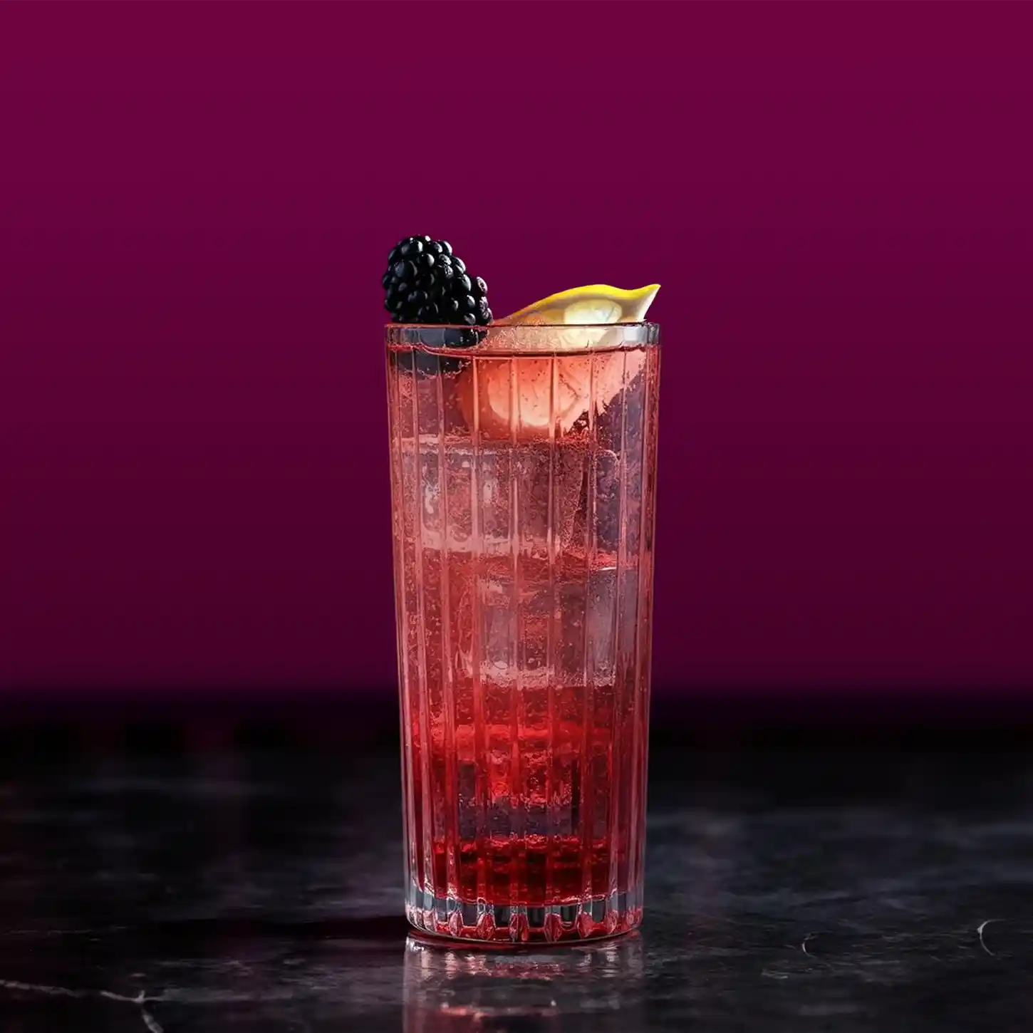 A tall glass filled with a red drink, ice cubes and garnished with a blackberry and a slice of lemon. The cup is on a dark, reflective surface with a dark purple curtain in the background.