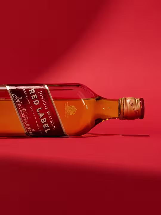 A bottle of Johnnie Walker Red Label whiskey lies on its side on a red background. Revealing the golden label and liquid, the bottle's cap is closed, and the lights highlight the curves and colours of the bottle.