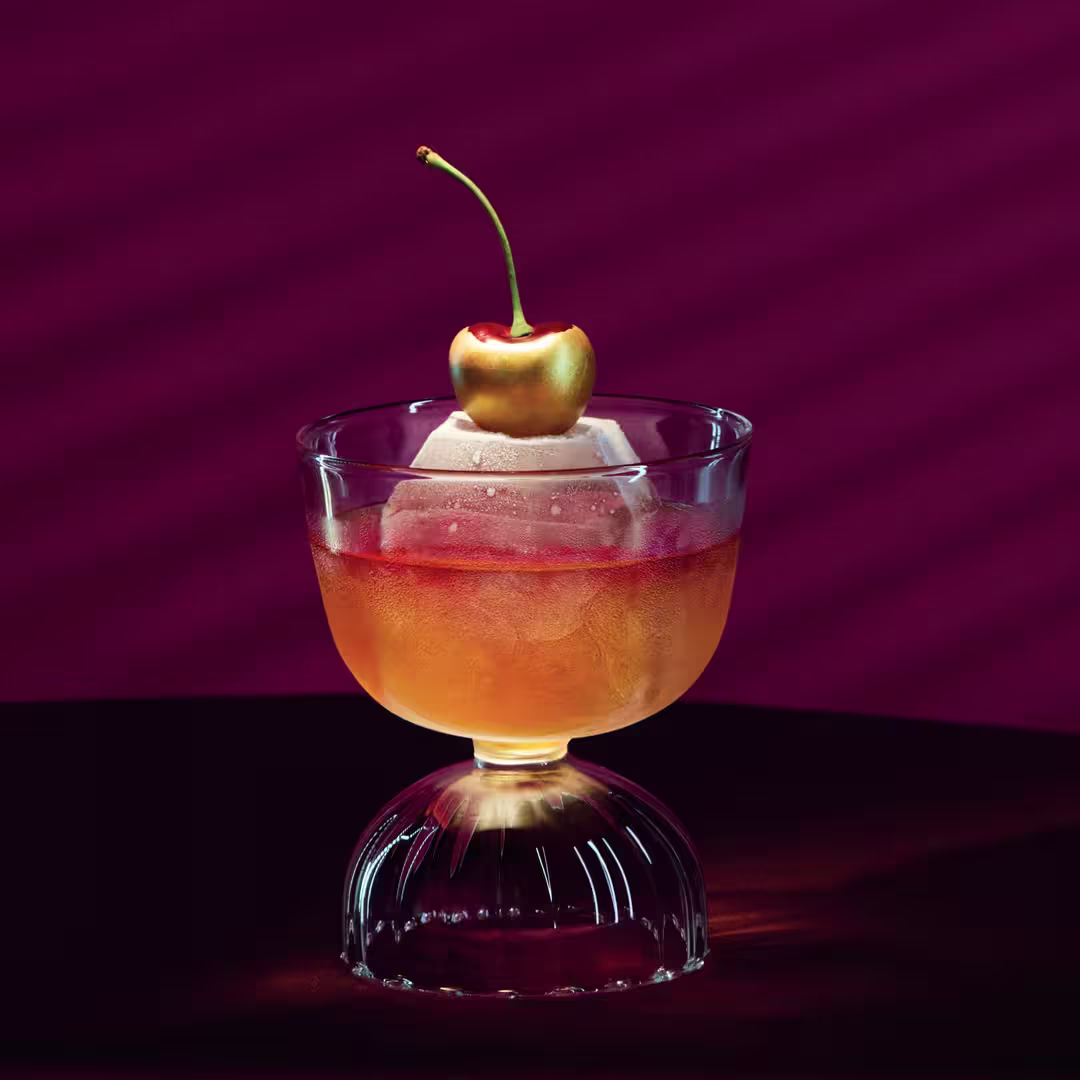 A cocktail in a glass with a large ice sphere and a cherry garnish sits on a table against a deep burgundy background with diagonal shadow lines.