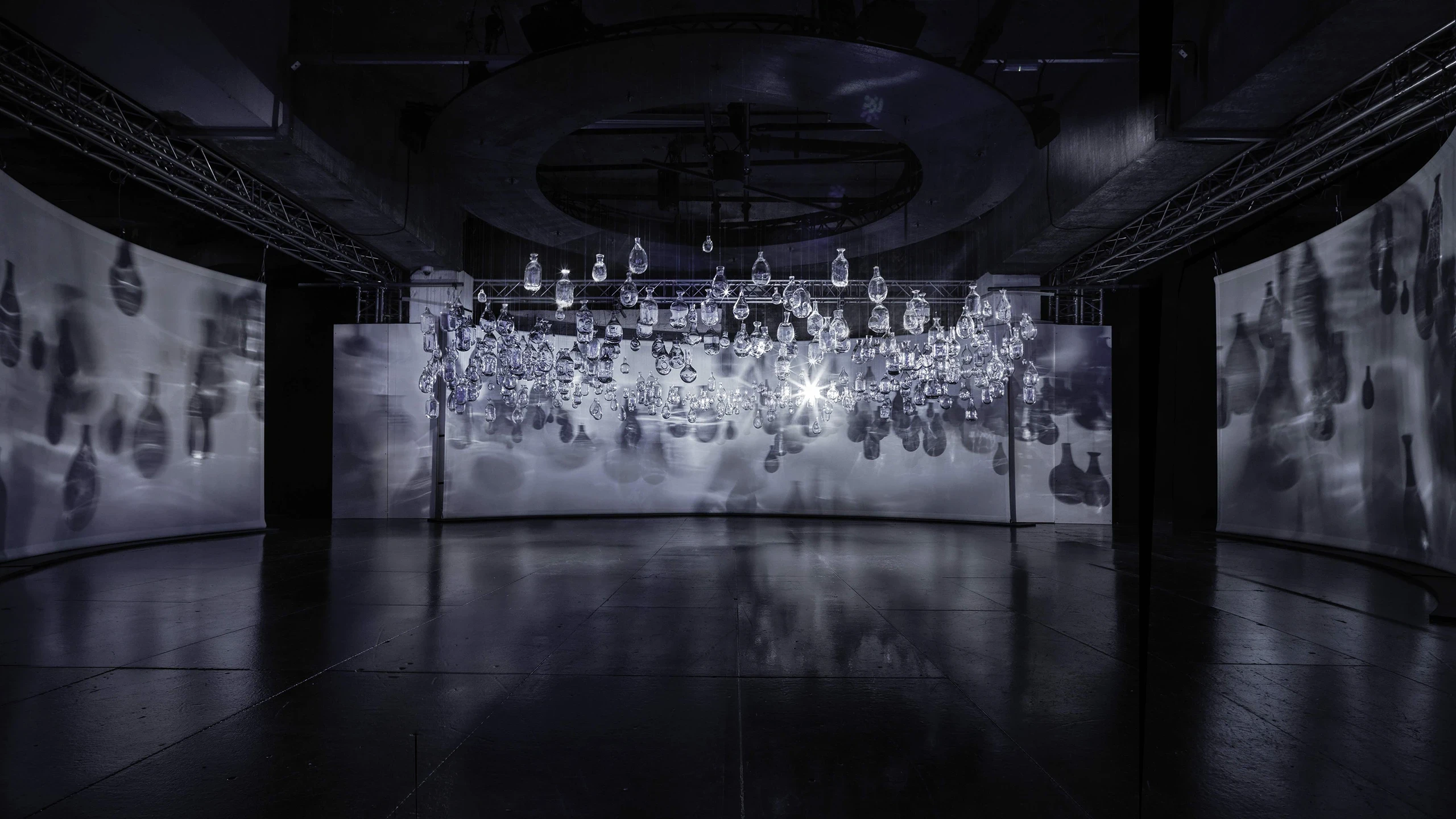 A dark, modern art installation features numerous hanging glass orbs reflecting and casting intricate shadows on curved white screens, with a bright light source in the center creating dramatic contrasts.