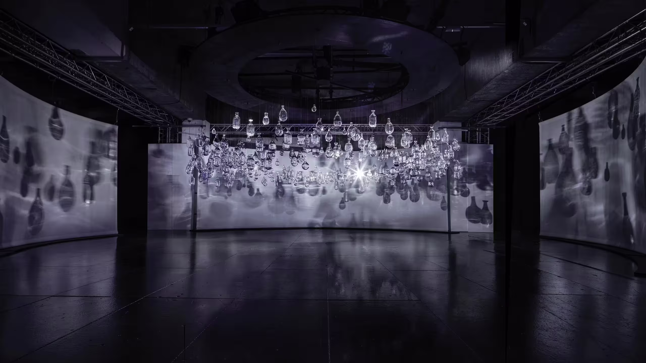Una habitación oscura y moderna cuenta con bombillas de vidrio colgantes suspendidas del techo, que proyectan sombras intrincadas sobre pantallas blancas curvas, con una fuente de luz brillante en el centro que crea reflejos dramáticos.