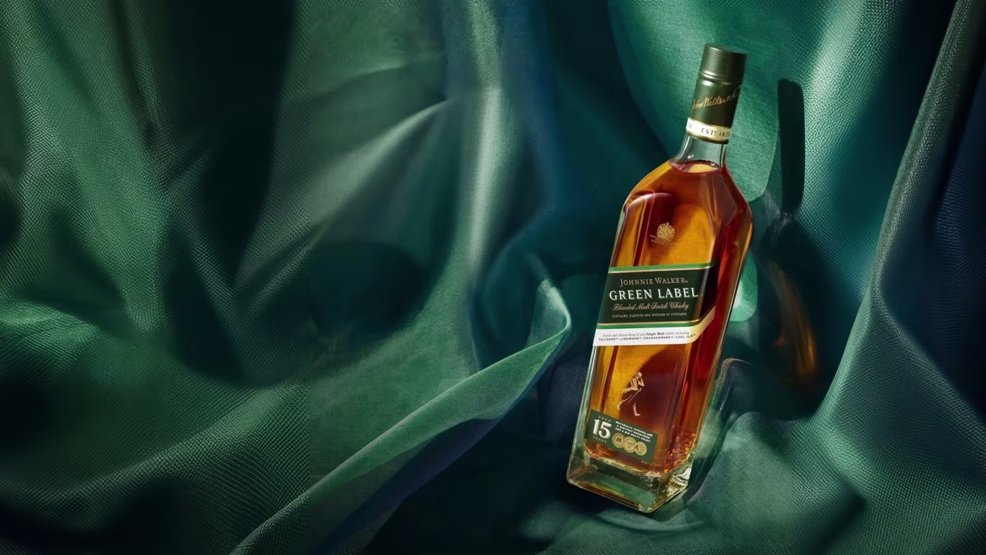 Una botella de whisky escocés de malta mezclado Johnnie Walker Green Label de 15 años se exhibe sobre una tela texturizada de color verde oscuro con una iluminación suave que resalta la botella.