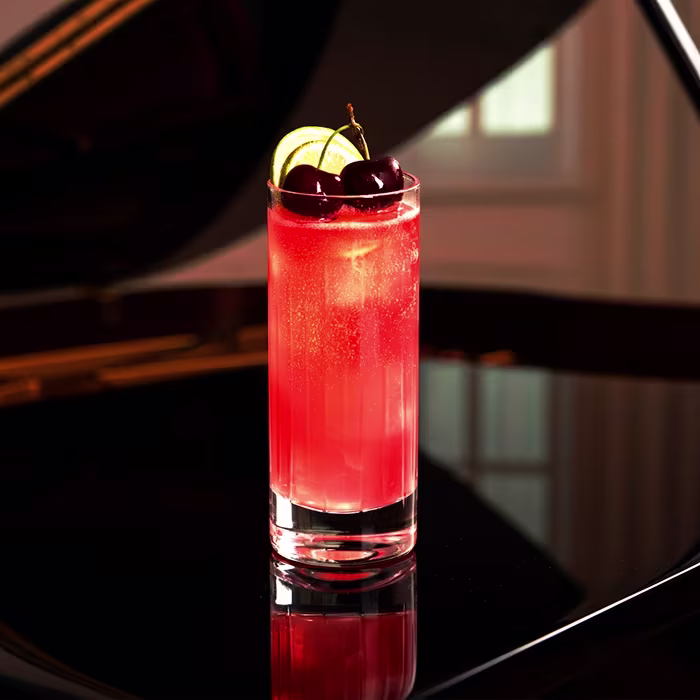  Un vaso alto de cóctel rojo brillante adornado con cerezas frescas y rodajas de lima reposa sobre un piano negro brillante, con una suave luz natural de fondo.