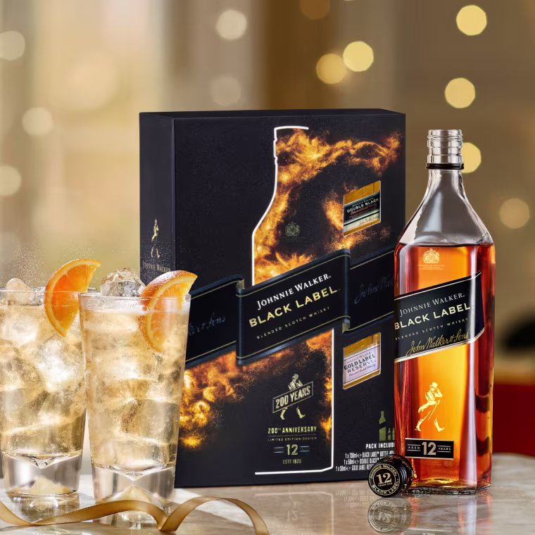 A bottle and box of Johnnie Walker Black Label 12 Year Old Scotch whisky beside two glasses with whisky, ice, orange slices, and a coin, all on a blurred golden background.