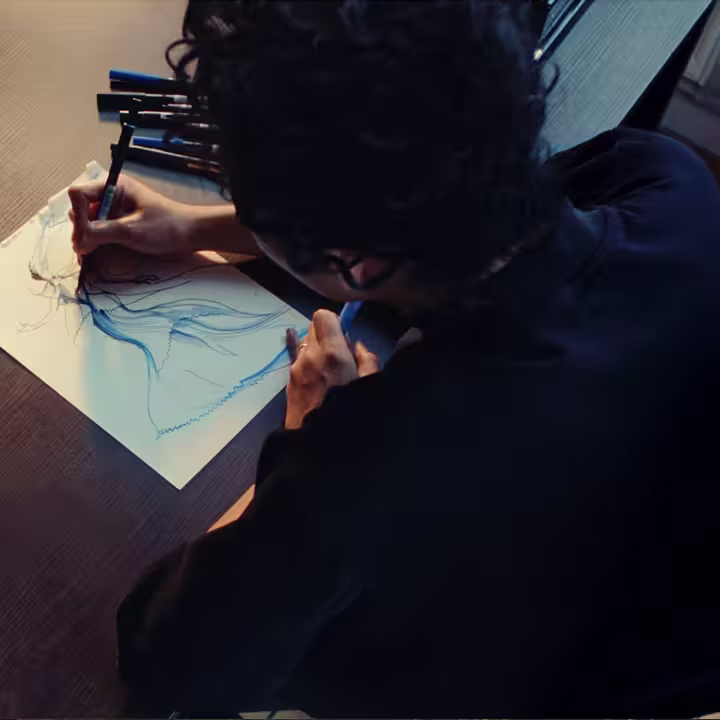 Robert Wun zit aan een tafel en tekent een gezicht met blauwe lijnen op wit papier. Verschillende stiften liggen verspreid over de tafel. De foto is van bovenaf genomen.