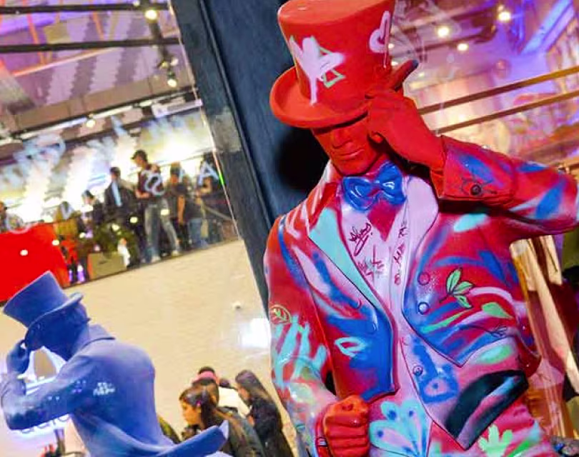 Dos coloridas estatuas de hombres con traje y sombrero de copa se encuentran en una vibrante galería. Una estatua es roja con un estampado blanco, y la otra, azul. Se ven personas a través de los grandes ventanales del fondo.