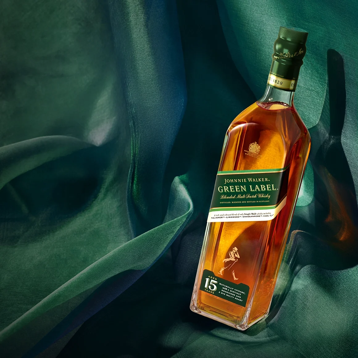 ขวดวิสกี้สก็อตช์มอลต์ผสม Johnnie Walker Green Label อายุ 15 ปี วางอยู่บนผ้าสีเขียวเข้มที่พลิ้วไหว พร้อมด้วยแสงไฟนวลๆ ที่เน้นให้ของเหลวสีเหลืองอำพันและฉลากสีเขียวของขวดโดดเด่นขึ้น