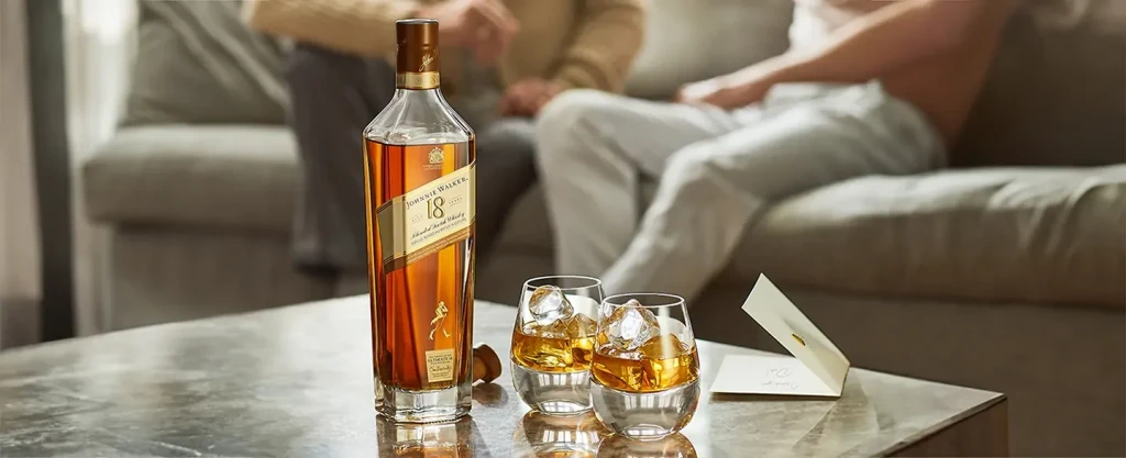 Una botella de whisky Johnnie Walker de 18 años y dos vasos con whisky y hielo se encuentran sobre una mesa, con dos personas sentadas en un sofá en el fondo borroso y un pequeño sobre sobre la mesa.