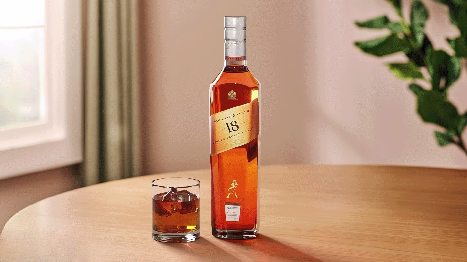 Ένα μπουκάλι Johnnie Walker 18 ετών blended σκωτσέζικο ουίσκι στέκεται σε ένα ξύλινο τραπέζι δίπλα σε ένα ποτήρι ουίσκι με πάγο. Υπάρχει ένα φυλλώδες πράσινο φυτό και ένα παράθυρο στο απαλά φωτισμένο φόντο.