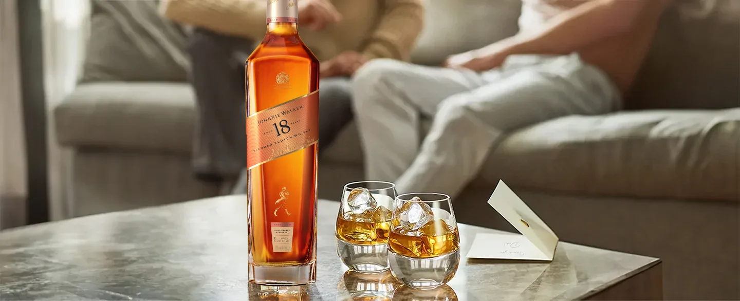 Một chai rượu whisky Johnnie Walker và hai chiếc ly đựng đầy rượu whisky và đá đặt trên bàn, với hai người ngồi trên ghế sofa ở phía sau mờ. Một tấm thiệp nhỏ nằm cạnh những chiếc ly.