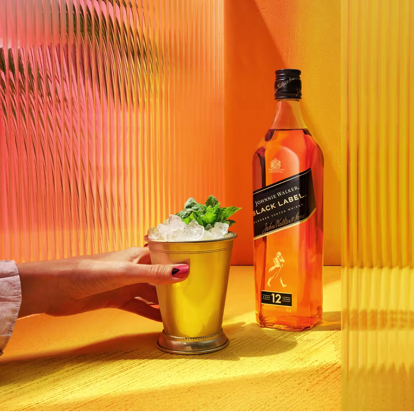 Бутилка Johnnie Walker Black Label стои върху жизнена жълта повърхност. До него ръка държи златна чаша, пълна с лед и листа от мента. Фонът включва цветни оранжеви и жълти текстурирани панели.