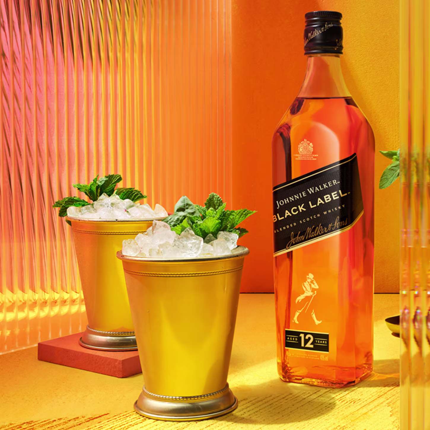 A hand holds a gold cup filled with ice and mint leaves next to a bottle of Johnnie Walker Black Label whiskey. The scene is set against a vibrant yellow and orange background, creating a warm, inviting atmosphere.