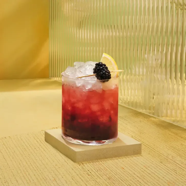 A cocktail in a glass filled with crushed ice sits on a square coaster. The drink is garnished with a blackberry and a lemon slice on a toothpick. The background features textured glass and a yellow surface.
