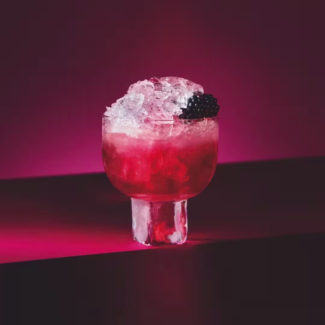 A glass of red cocktail filled with crushed ice and garnished with a blackberry, set against a deep red background with dramatic lighting.