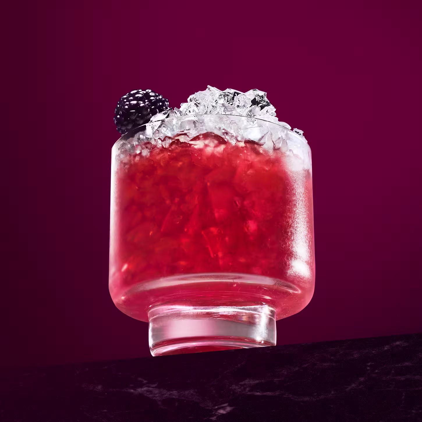 Een glas rode cocktail staat op een donker oppervlak, gevuld met gemalen ijs en versierd met een braambes erbovenop. De achtergrond is een rijke kastanjebruine kleur, die de levendige tint van de drank aanvult.