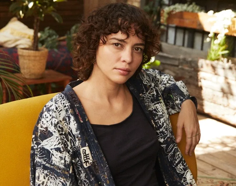 Pessoa com cabelo cacheado em uma camisa estampada senta-se em uma cadeira amarela. Uma planta é visível no fundo, adicionando um toque de verde ao cenário.