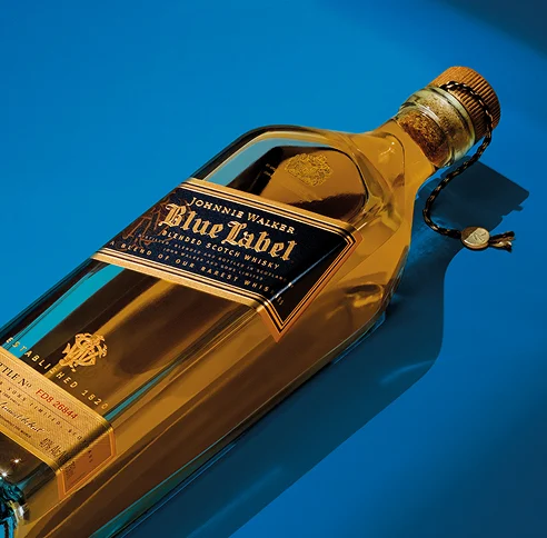 A bottle of Johnnie Walker Blue Label Scotch whisky lies on its side against a blue background. The bottle is transparent with a gold label and a wax seal hanging from its neck. The lighting highlights the amber color of the whisky.