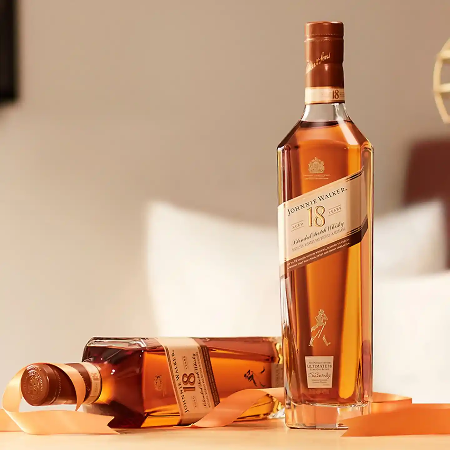 Johnnie Walker 18년산 스카치 위스키 두 병이 나무 표면에 진열되어 있습니다. 한 병은 똑바로 서 있고, 다른 한 병은 옆으로 누워 있고, 그 주위에는 금색 리본이 달려 있습니다. 배경은 부드럽게 흐릿하게 처리되어 있습니다.