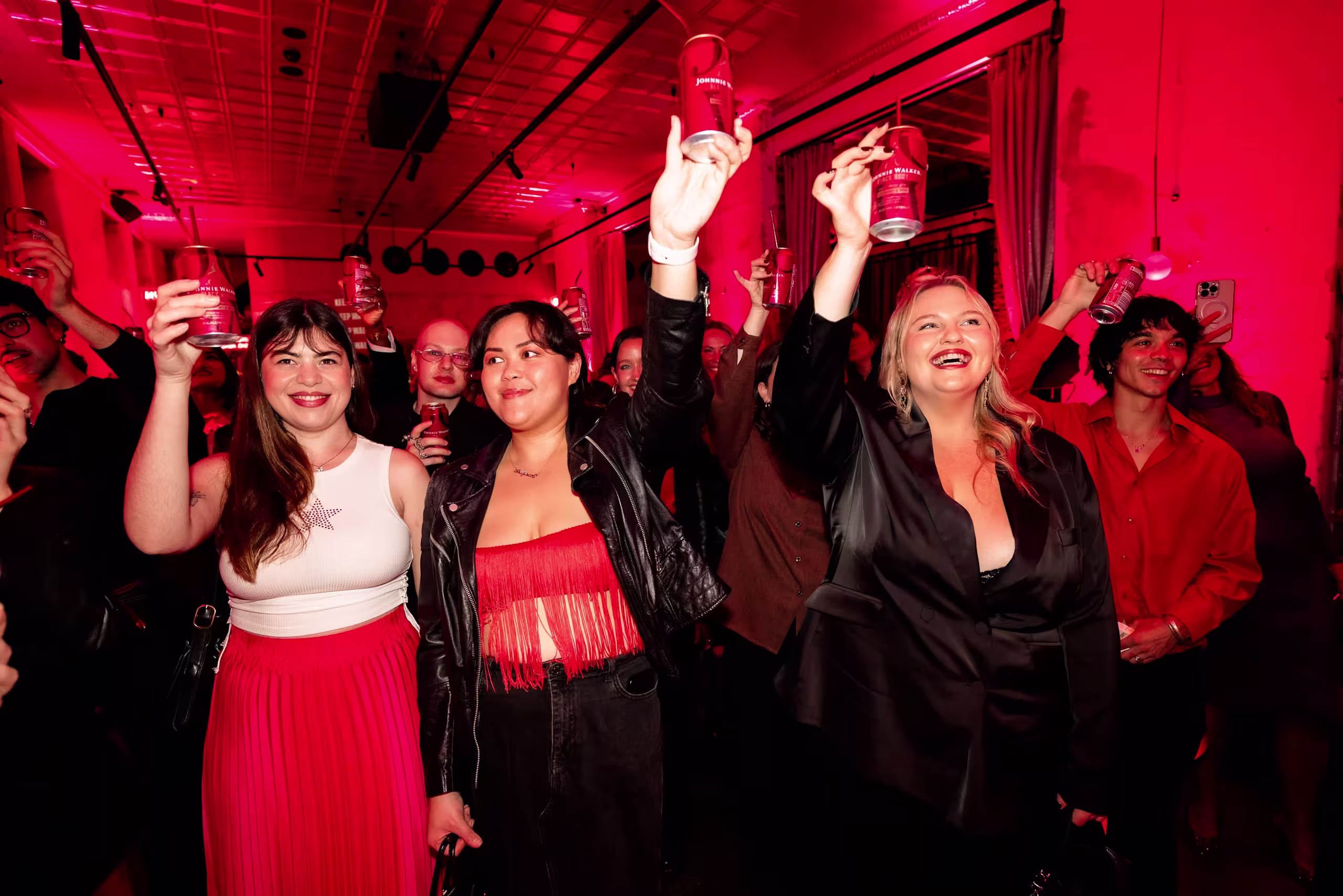 A group of people at a party raise their glasses in a toast, smiling and dressed in festive attire, with red lighting illuminating the room.