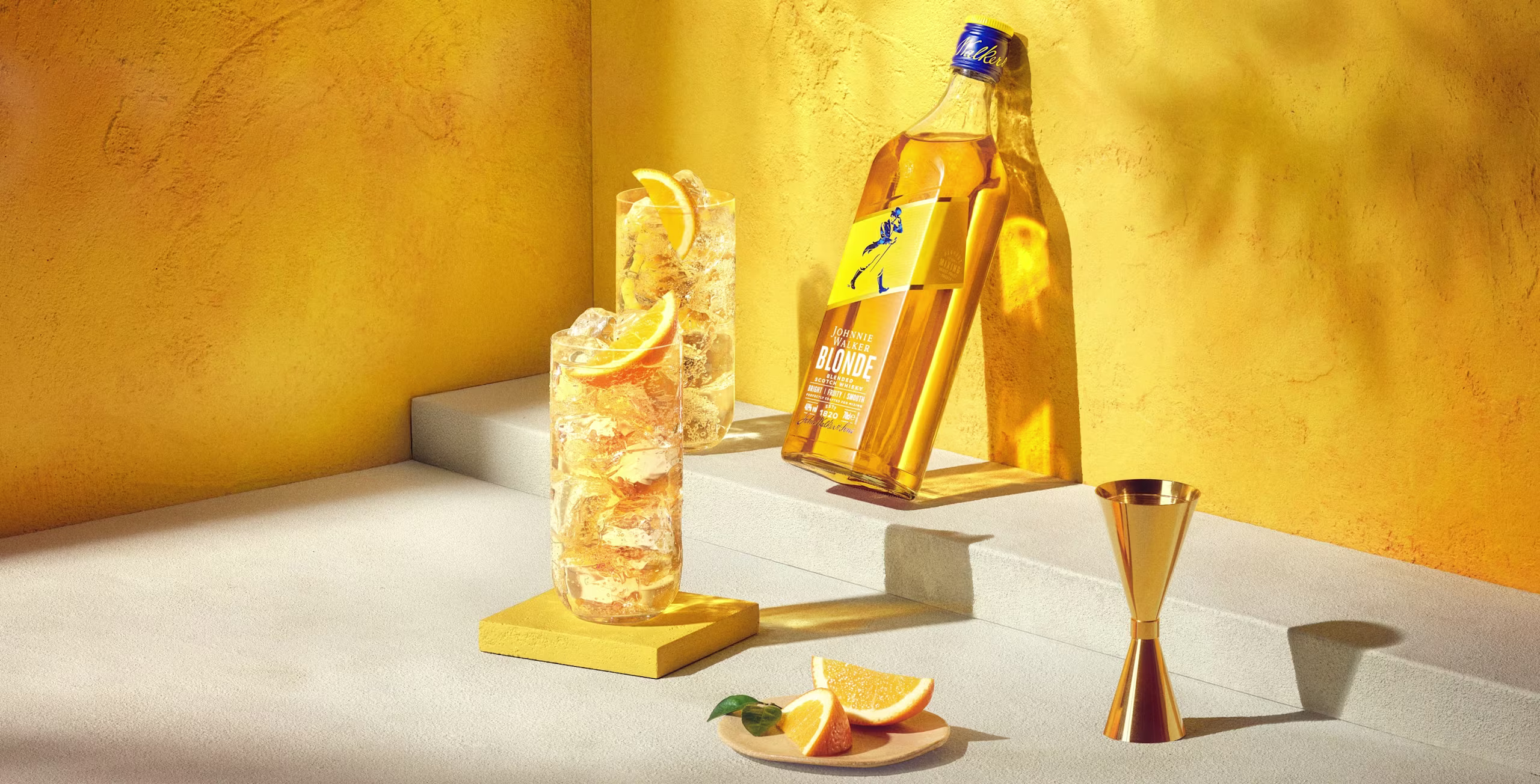 Butelka whisky z etykietą Johnnie Blonde stoi na słonecznej żółtej powierzchni. Obok niej szklanka wypełniona pomarańczowym napojem ozdobionym plasterkiem pomarańczy. Tło stanowi jasnożółta ściana z subtelnymi cieniami.