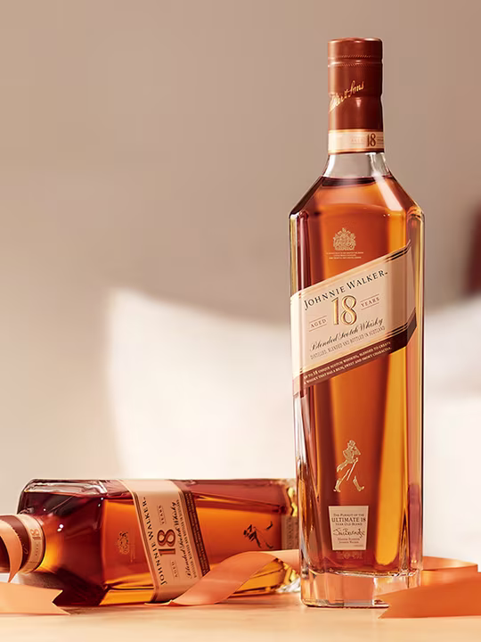 Two bottles of Johnnie Walker 18 year old blended Scotch whiskey are shown. One bottle stands upright, while the other sits on its side. Both are placed on a wooden surface, with soft, warm lighting highlighting the amber liquid.