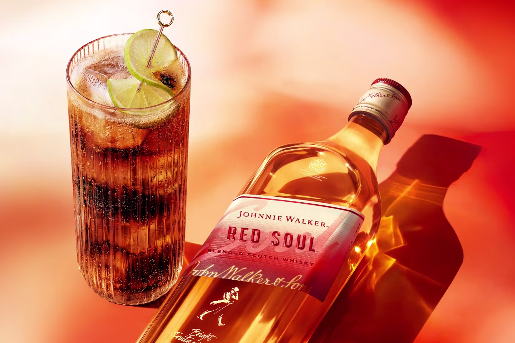 Butelka whisky Johnnie Walker Red Soul obok wysokiej szklanki wypełnionej ciemnym napojem, lodem i plasterkami limonki.