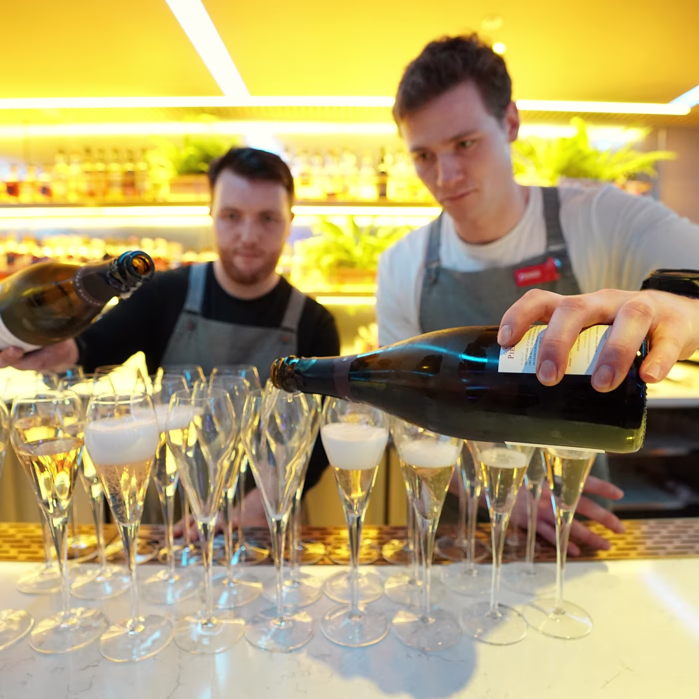 Two bartenders pour champagne into rows of glasses at a brightly lit bar.