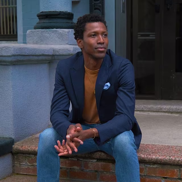 Un hombre con blazer azul marino, camisa marrón y vaqueros está sentado en una escalera exterior, mirando a un lado con las manos entrelazadas. Lleva un pañuelo azul en el bolsillo de su blazer. El fondo muestra parte de la entrada de un edificio.