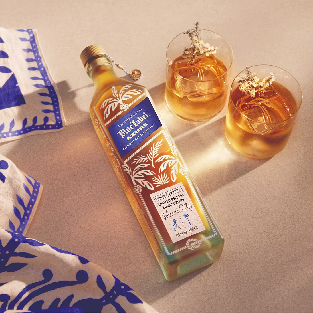 A bottle of Blue Label Azure whisky lies beside two glasses of whisky with ice and floral garnishes on a beige surface.