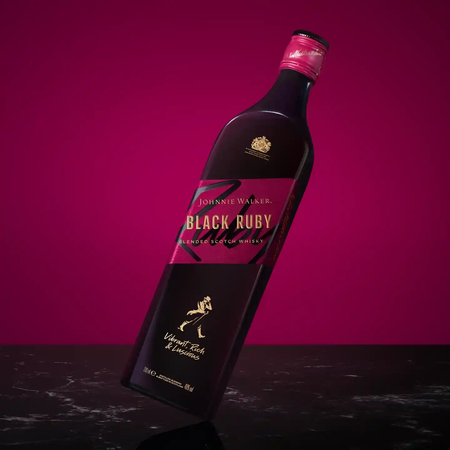 Una botella de whisky escocés mezclado Johnnie Walker Black Ruby sobre un fondo rosa oscuro. La etiqueta es negra con un detalle rosa y lleva el texto Black Ruby y detalles de la marca. La botella se encuentra inclinada sobre una superficie negra.