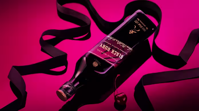 Una botella de whisky escocés mezclado Johnnie Walker Black Ruby sobre un fondo rosa oscuro. La etiqueta es negra con un detalle rosa y lleva el texto Black Ruby y detalles de la marca. La botella se encuentra inclinada sobre una superficie negra.