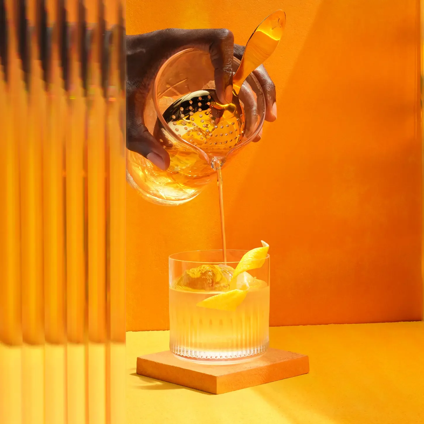 A hand pours a golden cocktail through a strainer into a glass with ice and lemon peel. The background is a vibrant orange, and a mirrored surface on the left reflects the scene.