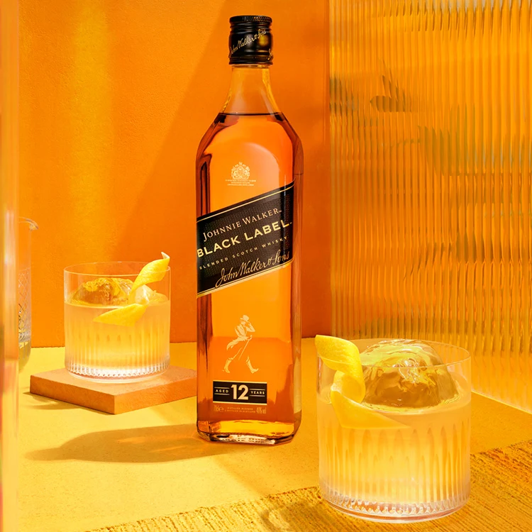 The Smooth & Floral Old Fashioned. A hand with dark skin pours a golden liquid through a strainer into a glass with ice and a lemon twist. The setting is vibrant orange with vertical reflections on the left. The glass is placed on an orange coaster.