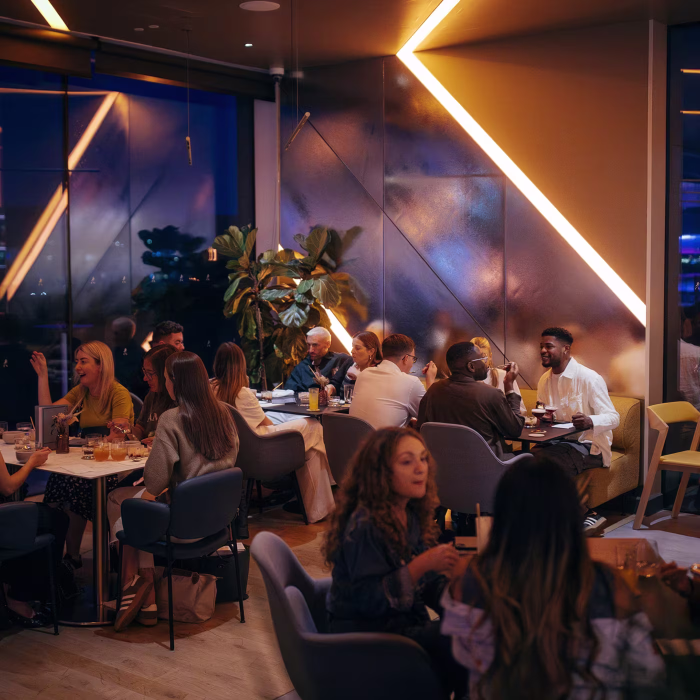 People dining and socializing in a modern restaurant with warm lighting and large windows at night.