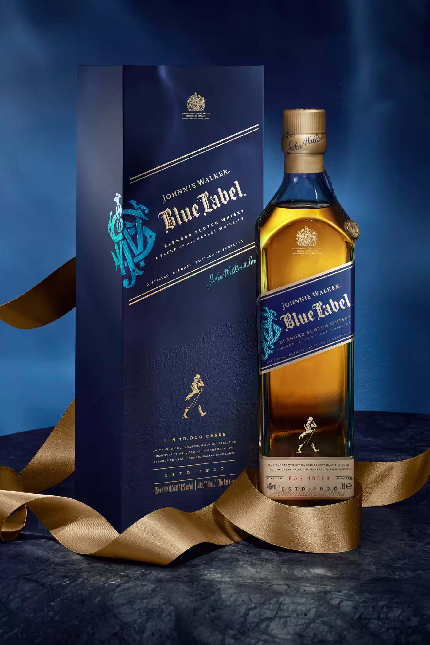 A bottle of Johnnie Walker Blue Label whisky stands next to its elegant blue box, both adorned with gold and blue accents; a gold ribbon is artfully draped around them on a dark marbled surface.