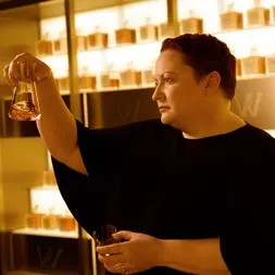 Dr. Emma Walker with short hair holds a glass vial, inspecting its contents, in a room filled with perfume bottles on shelves. The lighting is warm, creating an inviting ambiance. They are wearing a black outfit.