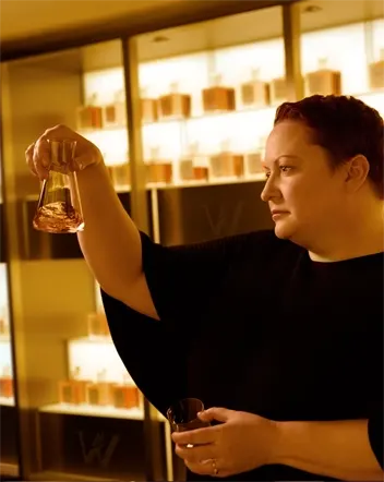 Dr. Emma Walker with short hair holds a glass vial, inspecting its contents, in a room filled with perfume bottles on shelves. The lighting is warm, creating an inviting ambiance. They are wearing a black outfit.