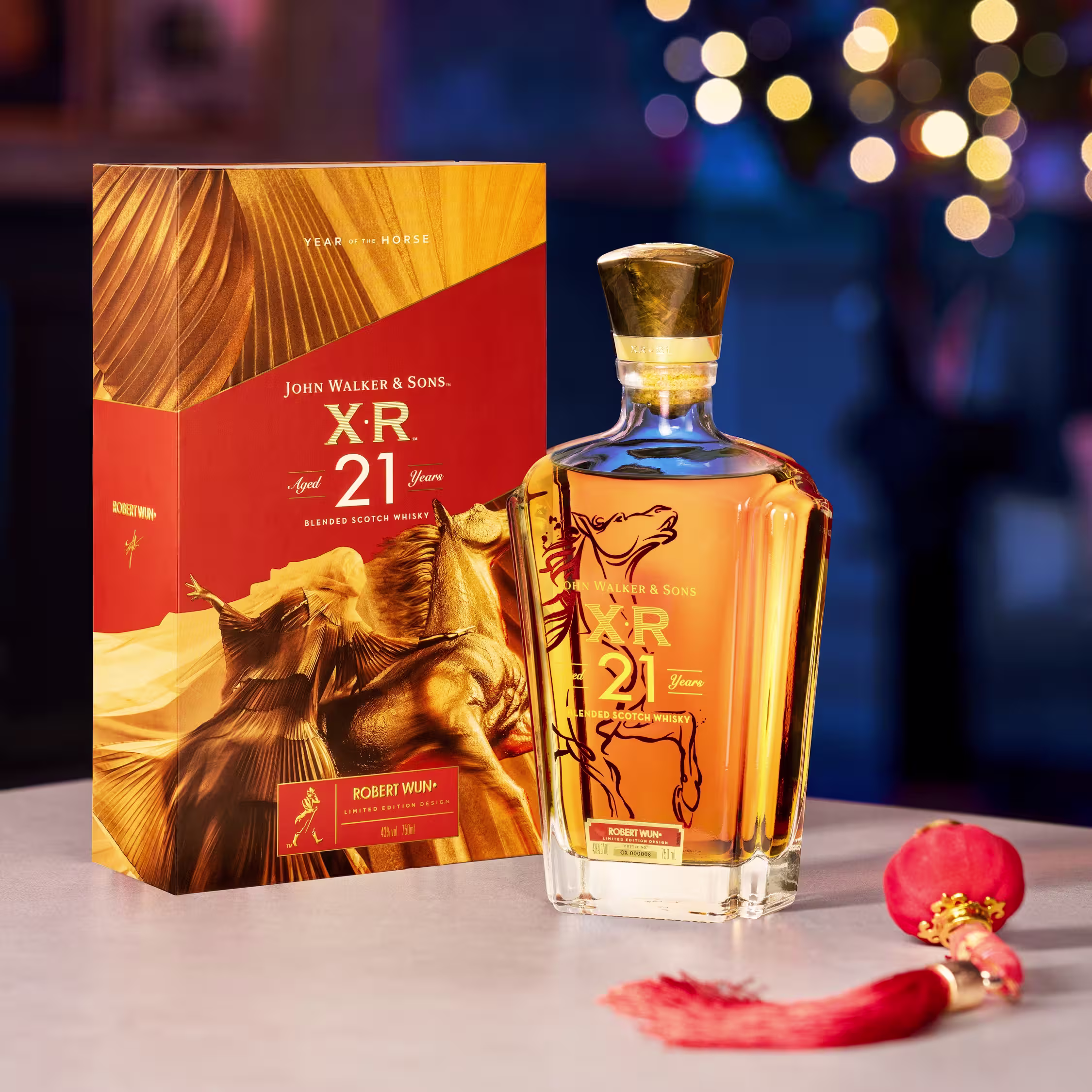 一瓶尊尼獲加 XR 21 年威士忌擺在桌上,旁邊是它的裝飾盒,附近有一個紅色流蘇裝飾品,背景是模糊的節日彩燈。