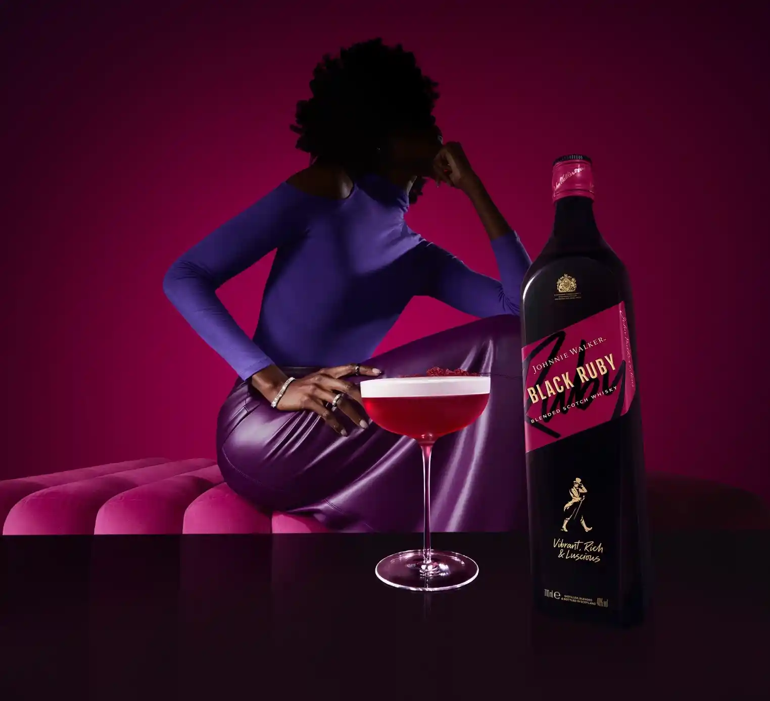 Una mujer con un vestido morado se sienta en un sofá rosa junto a un vaso de líquido rojo y una botella de Johnnie Walker Black Ruby.