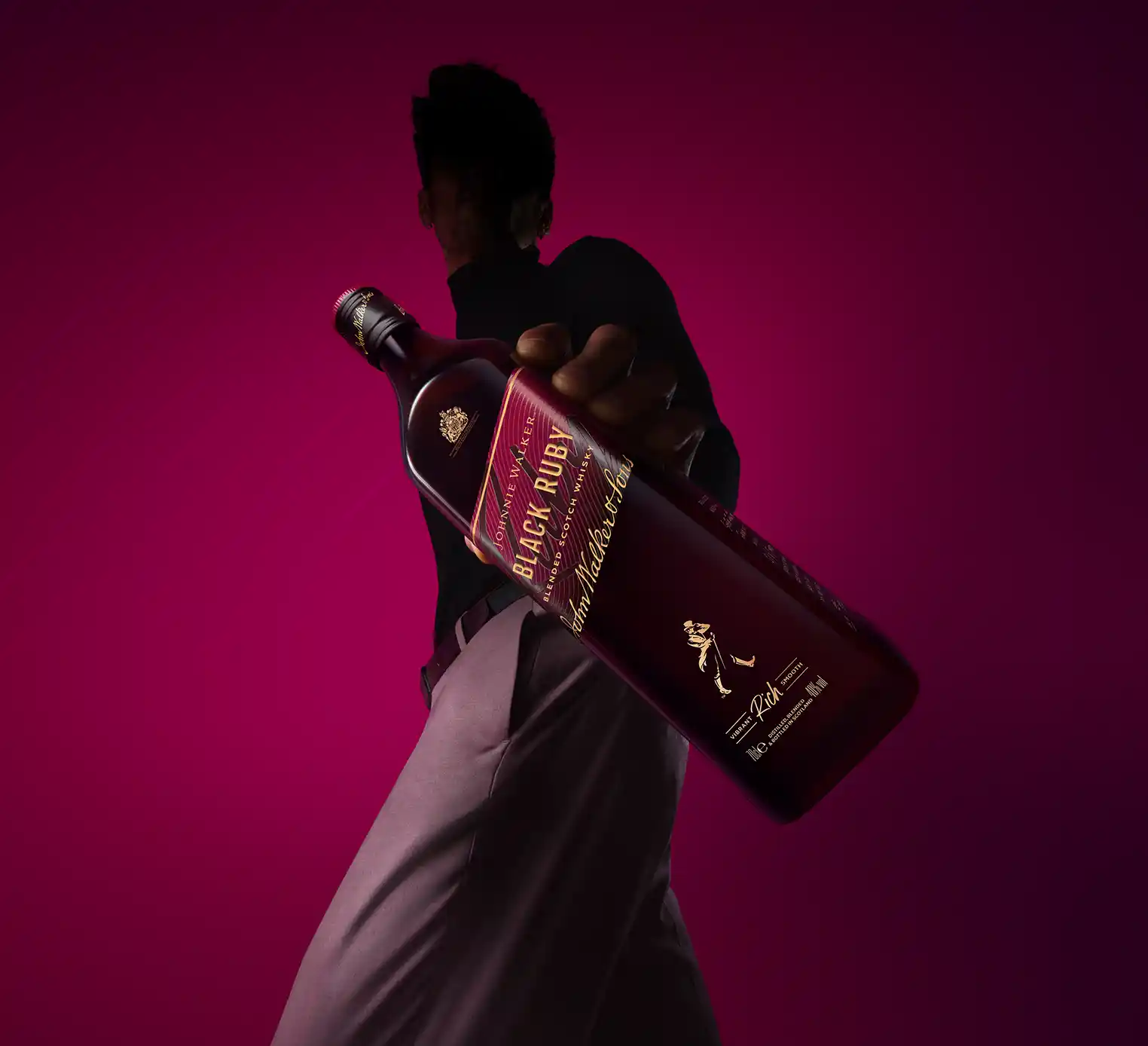 A person holds a large bottle of Black Label Scotch whiskey, tilted towards the camera. They are against a dark pink background, wearing a dark shirt and light pants. The image is taken from a low perspective.